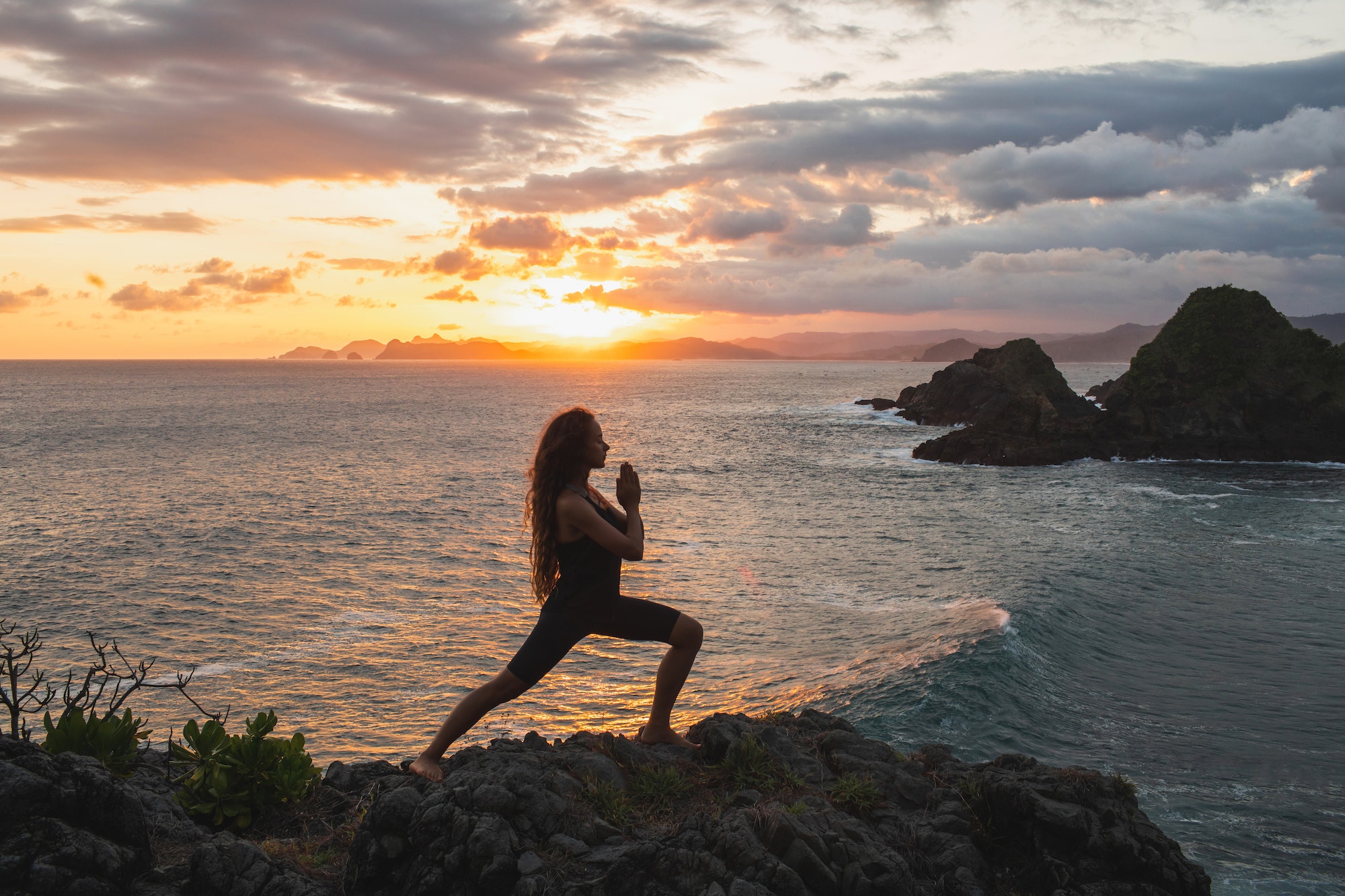 Young slim woman practicing yoga at sunset with beautiful ocean and mountain view.